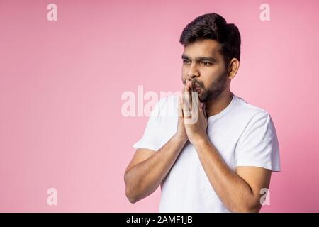 Closeup of young indian male in white t shirt, praying with palms clasped together, asking the God for good luck, success, health, on pink background Stock Photo