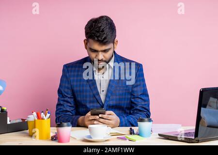 Stylish serious handsome young indian businessman sitting at desk in office, wearing checked suit, holding smartphone, checking email, using mobile ba Stock Photo