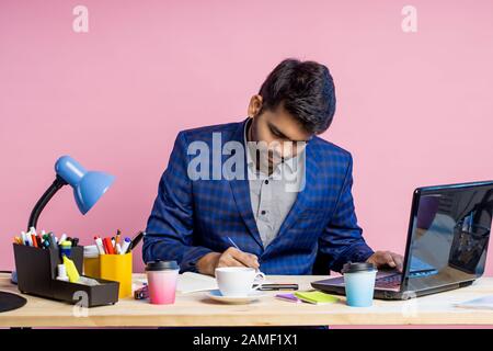 Serious confident young manager sitting at desk surrounded with stationery, dressed in suit, using laptop, writing something in notepad, making report Stock Photo