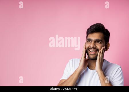 Portrait of pleasantly surprised happy man in white t shirt, touching cheeks with palms, looking aside with amazed expression, isolated on pink backgr Stock Photo