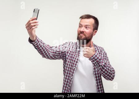 Like this phone! Portrait of excited handsome bearded man in plaid shirt taking selfie or talking on video call and showing thumbs up, satisfied with Stock Photo