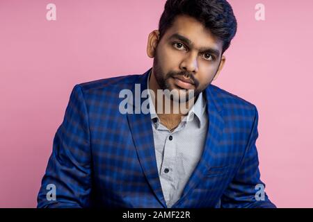 Horizontal closeup portrait of handsome confident businesslike young indian businessman sitting at workplace in office, wearing shirt, checkered suit, Stock Photo