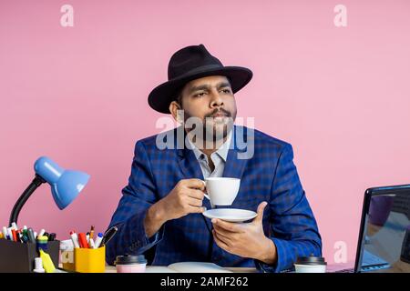 Photo of office worker, businessman in formal checkered suit, black hat , sitting at workplace, thinking about something, looking aside, enjoying arom Stock Photo
