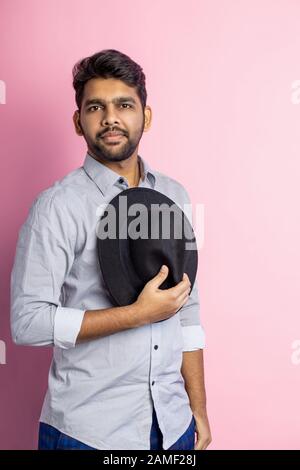 Vertical shot of confident serious calm young unshaven Indian man in gray shirt, with stylish haircut, holding black hat in his hand, looking at camer Stock Photo