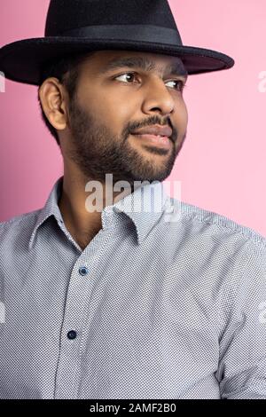 Vertical studio portrait of handsome unshaven young indian male, wearing gray shirt and black hat, looking to side with happy, satisfied expression, p Stock Photo