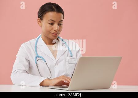 Portrait of beautiful female doctor using laptop while sitting against pale pink background, copy space Stock Photo