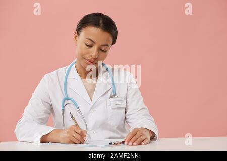 Portrait of beautiful female doctor writing on clipboard while filling in papers sitting against pale pink background, copy space Stock Photo