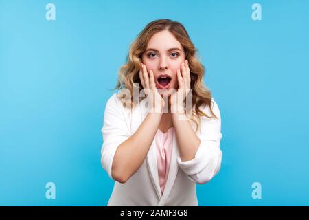 I can't believe this! Portrait of astonished woman with wavy hair in white jacket holding face and keeping mouth open, shocked by unexpected crazy new Stock Photo