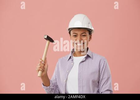 Waist up portrait of pretty female worker holding hammer and smiling happily while standing against pale pink background, copy space Stock Photo