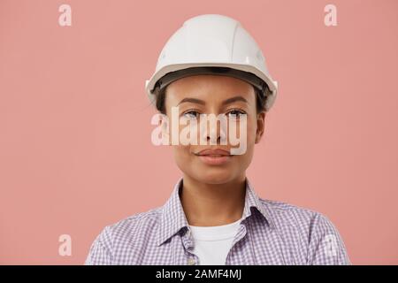 Head and shoulders portrait of beautiful female worker wearing hardhat and looking at camera while posing confidently against pale pink background, copy space Stock Photo