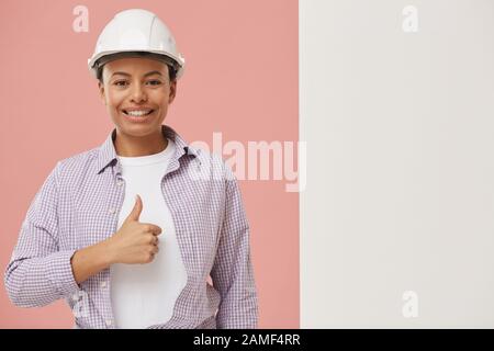 Waist up portrait of beautiful female worker showing thumbs up and looking at camera while standing by blank white sign against pale pink background, copy space Stock Photo