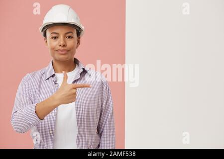 Waist up portrait of beautiful female worker looking at camera and pointing at blank white sign against pale pink background, copy space Stock Photo