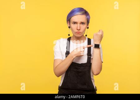 Portrait of dissatisfied angry hipster girl with violet hair in denim overalls pointing watch on her wrist and looking impatient, meaning no time, too Stock Photo