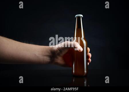 Men holding cold unopened bottle of beer with cap on black background. Hand holding glass of refrigerated wheat or lager beer on dark background Stock Photo