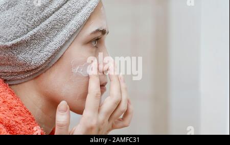 Young beautiful girl smearing her face with homemade natural cosmetic mask and looking in the mirror. Skin care and health care concept. Place for you Stock Photo