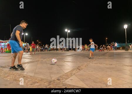 Naxos Greece - August 11 2019; Boy and father play football in square at night with focus on boy. Stock Photo