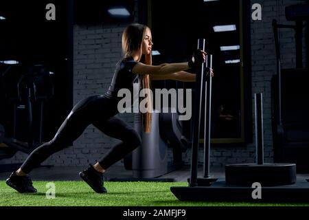 Side view of fit young woman in black sports outfit moving stand with weights in gym. Strong attractive female athlete with long hair pushing large equipment. Concept of bodybuilding, workout. Stock Photo