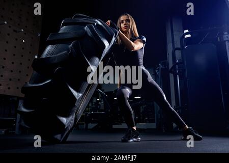 Front view of fit young woman in black sports outfit moving giant tire in gym. Strong attractive female athlete with long hair pushing large wheel. Concept of bodybuilding, workout. Stock Photo