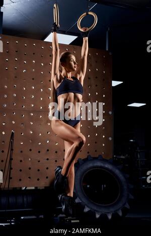 Front view of young attractive sportswoman doing ring exercise at gym. Fit female posing, doing fitness workout complex using gymnastic rings. Concept of sport, gymnastic. Stock Photo