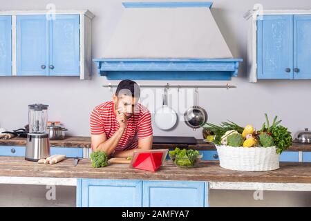 Thoughtful confused man watching food blog using tablet in modern kitchen, looking for dish recipe on internet to cook vegetarian salad with green fre Stock Photo