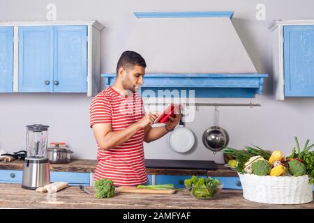 Serious attentive young man watching food blog using tablet in modern kitchen, searching dish recipe on internet to cook vegetarian salad with green f Stock Photo