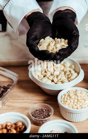 cropped view of chocolatier in black latex gloves holding white chocolate chips Stock Photo