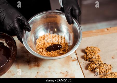 cropped view of chocolatier in black latex gloves holding bowl with flakes near delicious chocolate candy Stock Photo
