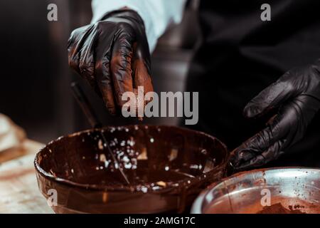 cropped view of chocolatier in black latex glove holding chocolate ball near bowl with melted chocolate Stock Photo