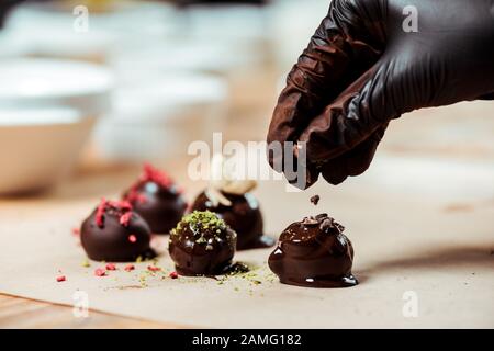 cropped view of chocolatier in black latex glove adding chocolate shavings on fresh made candies Stock Photo