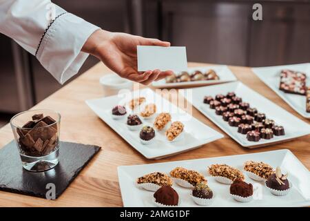 cropped view of chocolatier holding blank card in hand near tasty chocolate candies on plates Stock Photo