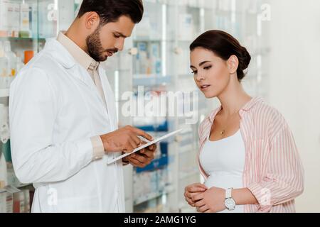 Druggist using digital tablet beside pregnant customer with medicaments on shelves at background Stock Photo