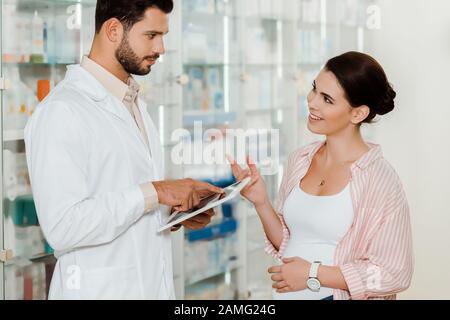 Side view of pharmacist with digital tablet looking at smiling pregnant woman in drugstore Stock Photo