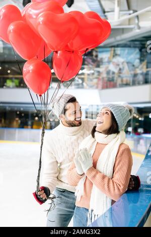 excited couple in winter outfit with red heart shaped balloons spending time on skating rink on valentines day Stock Photo