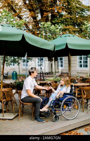 Young pretty woman in wheelchair spending time together with male boyfriend, holding hands, at outdoor cafe in the old city center Stock Photo