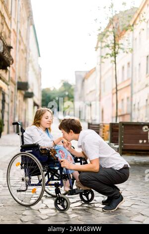 Handsome happy man smiling and holding hand of his beautiful beloved blond handicapped woman in wheelchair while walking together on the street of old Stock Photo