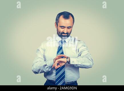 Serious adult bearded man in elegant white shirt with blue tie looking to his watch isolated on light green yellow background. Successful entrepreneur Stock Photo