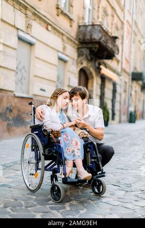 Young man hugs his pretty woman on the wheelchair, while walking together on the street of city. Couple in love in wheelchair embracing and touching Stock Photo