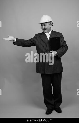 Studio shot of mature Asian businessman as engineer with hardhat against gray background in black and white Stock Photo