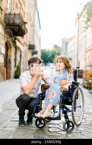 Young lovely Couple In Wheelchair, Enjoying A Day In The City, handsome man holding and kissing the hand of pretty young woman in wheelchair Stock Photo