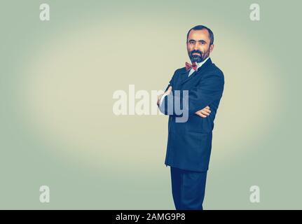 Serious bearded man in dark suit, blue shirt, red bow tie, looking to the camera on green-yellow background. Successful, confident boss. Elegant manag Stock Photo