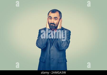 Astonished, bearded businessman covering his ears with hand palms isolated on light green yellow wall background. Facial expressions, feelings, body l Stock Photo