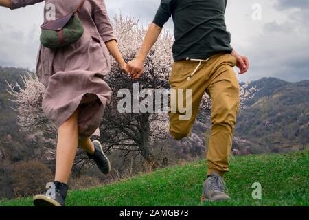 A guy with a girl run holding hands among the flowering trees outdoors. Spring time. A girl with long hair. On the girl a long linen dress Stock Photo