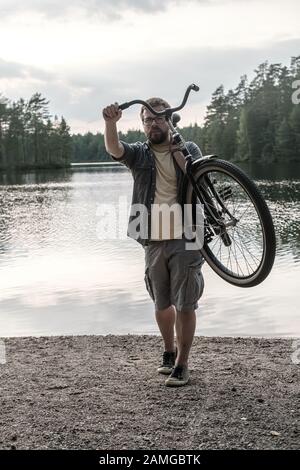 Bearded man put a bicycle on shoulder and carries it, from a sandy beach, against the background of a forest lake and cloudy sky. Stock Photo