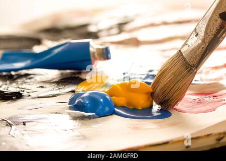 Artist brush with paint from painting tubes ready to mix on a palette. Blue color tube in the background. Backlit close-up image with selective focus. Stock Photo