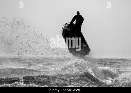 Dramatic, monochrome action shot of isolated male on jet ski bike in sea, in silhouette, flying high in the air, sea spray, waves below. Stock Photo