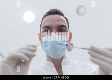 Dentist wearing surgical mask while holding angled mirror and drill, ready to begin Stock Photo