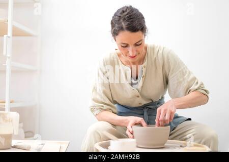 Young female sitting by table and making clay or ceramic mug in her working studio Stock Photo