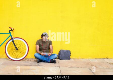 Cheerful handsome bearded man using a mobile phone while listening to music in headphones, spending time with pleasure while sitting near the bike, ou Stock Photo