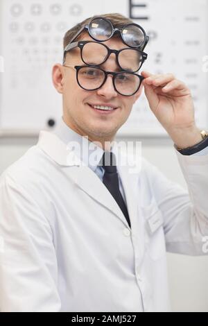 Portrait of smiling young ophthalmologist wearing several glasses while posing in office next to visual chart, copy space Stock Photo