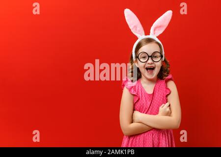 Happy easter. Funny cheerful little girl in rabbit ears and glasses on a red background. The child crossed his arms and looks at the camera. Stock Photo